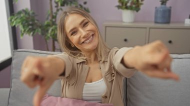 Güzel, sarışın, genç bir kadın kapalı bir oturma odasında gülümsüyor, arka planda bitkilerle neşeyle işaret ediyor..