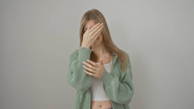 Yeşil kazaklı genç sarışın bir kadın beyaz bir arka plana karşı elleriyle gözlerini kapatıyor.