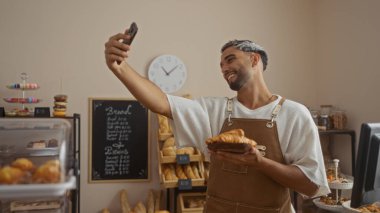 Genç, Arap, sakallı bir adam arka planda ekmek ve hamur işleriyle dolu raflarla kruvasan tutarken fırında neşeyle selfie çeker.
