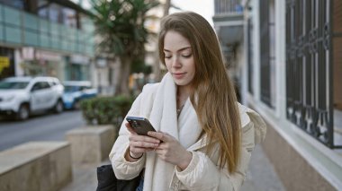Odaklanmış beyaz bir kadının portresi, arka planda arabalar ve binalar olan bir caddede dikilirken akıllı telefonunu kullanıyor..