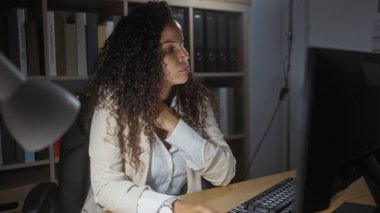 Ofisteki kıvırcık saçlı, boynu ağrıyan genç İspanyol kadın. Profesyonel bir şekilde bilgisayar ekranına bakarken boynu ağrıyor.