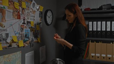 Genç, kızıl saçlı bir kadın loş bir dedektifin ofisindeki mantar panoda ipuçlarını analiz ediyor.