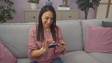 Gülümseyen genç bir kadın, rahat bir oturma odasında, akıllı telefonuyla koltukta oturur ve sohbet eder.