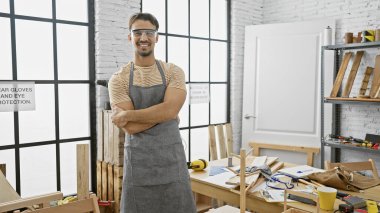 Sakallı ve kollarını kavuşturmuş İspanyol bir adam koruyucu gözlük ve önlük takarak bir marangozluk atölyesinde duruyor..