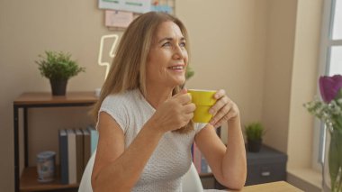 Gülümseyen olgun bir kadın rahat bir oturma odasında kahveden zevk alır, rahat bir yaşam tarzını ve ev rahatlığını somutlaştırır..