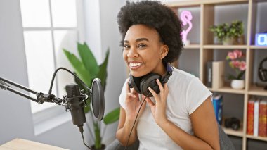 Radyo stüdyosunda gülümseyen Afrikalı Amerikalı bir kadın kulaklık takıyor ve mikrofonun yanında oturuyor..