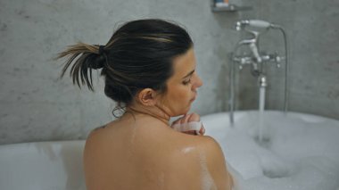 Genç, yetişkin bir esmer kadın köpüklü bir ev banyosunda dinleniyor. Huzur ve kendine bakıcılık yayıyor..