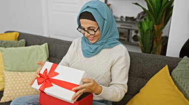 Başörtüsü takan gülümseyen bir kadın renkli yastıklarla çevrili rahat bir kanepede hediye kutusunu açar..