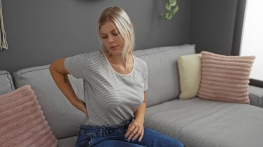 Modern oturma odasında oturan genç bir kadın, belini acıdan tutarak, sarı saçlar ve rahat yastıklı, şık gri bir iç mekâna karşı günlük giysiler giyiyor..