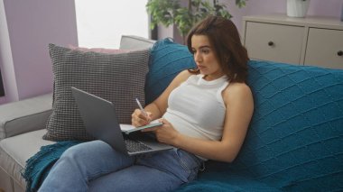 Esmer saçlı İspanyol bir kadın oturma odasında oturuyor, dizüstü bilgisayarla çalışıyor ve bir deftere yazı yazıyor..