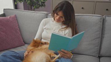 Genç bir kadın evinde rahatlıyor, kanepede uyuyan Pomeranian köpeğiyle kitap okuyor, evde rahat bir sahne tasvir ediyor..