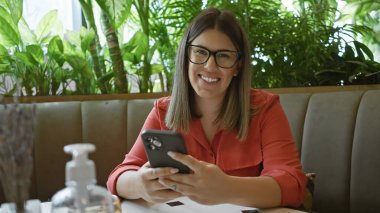 Akıllı telefon kullanan, gözlüklü, gülümseyen genç bir kadın, etrafı bitkilerle çevrili bir kafe masasında oturuyor..