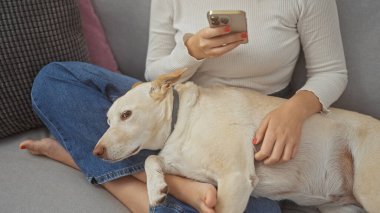 Genç bir kadın, rahat bir oturma odasında köpekle kucaklaşırken akıllı telefonunu kullanarak kanepede uzanıyor..