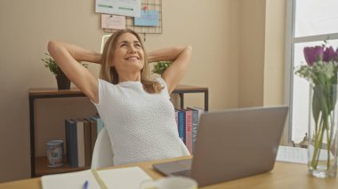 Elleri arkada, ofiste dizüstü bilgisayar, kitap ve çiçeklerle rahatlayan olgun bir kadın.