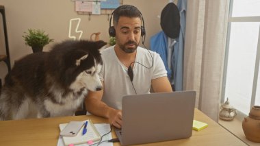 Beyaz tişörtlü ve kulaklıklı genç bir İspanyol adam rahat bir ofis ortamında köpeğinin yanında dizüstü bilgisayarda çalışıyor..