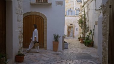 Genç İspanyol bir kadın polignano 'nun büyüleyici sokaklarında yürüyor. Bir kısrak, puglia, İtalya, Avrupa. Etrafında tuhaf taş binalar ve saksı bitkileri var..