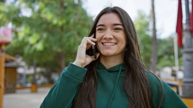 Gülümseyen genç bir İspanyol kadın güneşli bir şehir parkında telefonla sohbet ediyor..