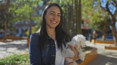 Güneşli bir şehir parkında elinde bichon köpeğiyle gülümseyen bir kadın.