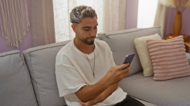 Sakallı genç bir adam oturma odasında oturuyor, akıllı telefonunu kapalı bir dairede kullanıyor..