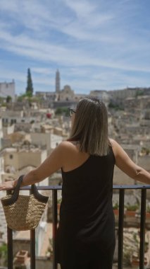 Genç bir İspanyol kadın, siyah bir gömlek giymiş ve elinde dokuma çantasıyla, Materya, Basilicata, İtalya 'nın güzel bir bakış açısıyla eski kasabayı seyrediyor..