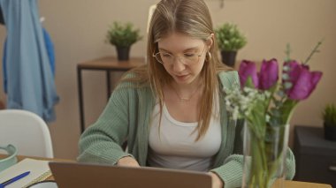 Gözlük takan odaklanmış genç bir kadın rahat bir ev ortamında dizüstü bilgisayarda çalışıyor. Yanında çiçekler ve bitkiler var..