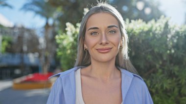 Açık hava parkında yeşil yapraklı sarı saçlı, gülümseyen beyaz bir kadının portresi.