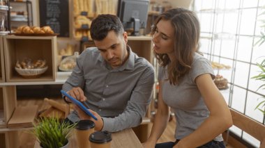 Bir çift fırında otururken tablete bakıyor, arka planda ekmek ve hamur işleri, masada iki kahve fincanı, ev ortamında, kadın izlerken erkek ekranı işaret ediyor.
