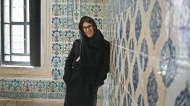 Gözlüklü ve siyah ceketli gülümseyen genç bir kadın tarihi İstanbul Sarayı 'nda süslü döşemelerin yanında duruyor.