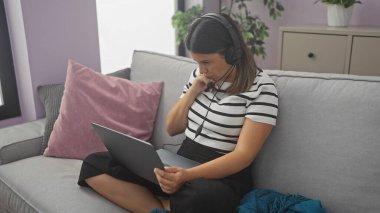 Kulaklık takan odaklanmış bir kadın koltukta otururken dizüstü bilgisayar kullanıyor..