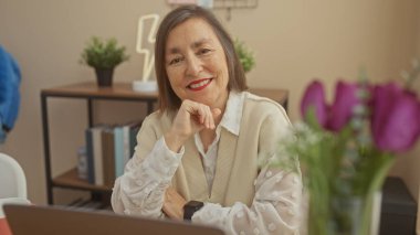 Gülümseyen orta yaşlı, kısa saçlı bir kadın modern dekorlu, rahat bir odada oturuyor, dizüstü bilgisayarını canlı çiçeklerin yanında kullanıyor..