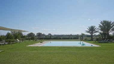 Parlak mavi bir gökyüzünün altında İtalya 'nın Puglia kırsalında yemyeşil bahçeler ve palmiyelerle çevrili sakin bir açık yüzme havuzu..