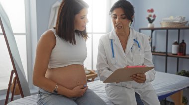 Hamile bir kadın klinikte kadın doktora danışarak hamilelik ve tıbbi bakımı tartışıyor..