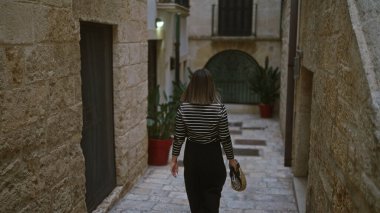 Polignano 'da dar taştan caddelerde yürüyen genç İspanyol kadın bir kısrak, puglia, İtalya, şık bir saman çantası tutuyor..