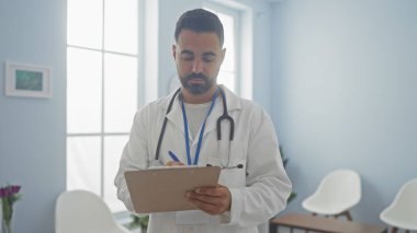 A focused hispanic man wearing a lab coat and stethoscope takes notes in a bright clinic room. clipart