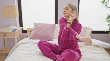 Pembe pijamalı genç bir beyaz kadın bir yatakta oturuyor, modern bir yatak odasında uzanıyor..