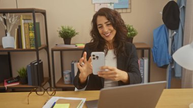 Modern bir ev ofisinde akıllı telefon kullanan gülümseyen bir kadın dizüstü bilgisayarı, gözlüğü ve kapalı bitkileri olan bir kadın..