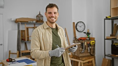 Yakışıklı, sakallı, ahşap atölyesinde elinde bir pano tutarken gülümseyen bir İspanyol.