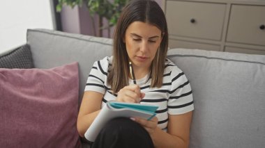 Gri bir kanepede oturan düşünceli genç bir kadın modern bir oturma odasındaki deftere yazıyor..