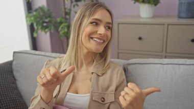 Sarışın, güzel bir kadın. Oturma odasında oturuyor. Gülümsüyor ve parmaklarıyla işaret ediyor..