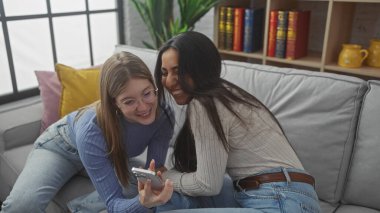 İki kadın gülüyor ve arka planda kitaplar olan bir kanepede birlikte akıllı telefona bakıyorlar.