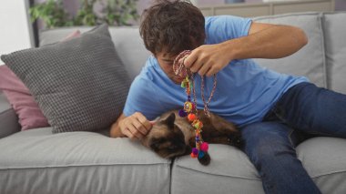 Genç bir İspanyol adam, içeride kanepede siyam kedisiyle oynuyor..