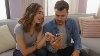 Kadın ve erkek evdeki kanepede oturuyor, rahat oturma odasında cep telefonlarına bakıp gülümsüyorlar.