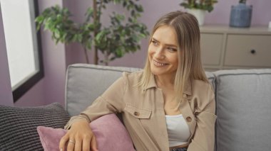 Güzel, sarışın, beyaz bir kadın gri bir kanepede otururken gülümsüyor rahat bir oturma odasında bitkiler ve pastel mor duvarlarla..