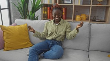 Gülümseyen Afrikalı Amerikalı kadın rahat bir oturma odasında gri bir kanepede dinleniyor.