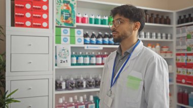 Beyaz eczacı önlüklü sakallı bir genç düşünceli bir şekilde kapalı bir eczanede duruyor..