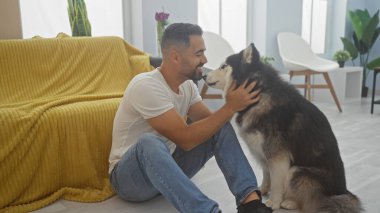 Gülümseyen genç İspanyol bir adam, sıcak, aydınlık, modern bir evin içinde köpeğini sevgiyle okşuyor..