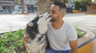 Genç bir İspanyol erkeği, şehir parkında evcil köpeği Husky 'nin yüzünü sevgiyle yalıyor.