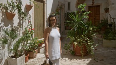 Beyaz elbiseli güzel, genç bir İspanyol kadın Locorotondo, Puglia, İtalya 'nın büyüleyici eski caddelerinde yürüyor. Etrafı yemyeşil ve tarihi mimariyle çevrili..