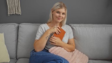 Oturma odasındaki kanepede oturan genç sarışın kadın, elinde bir kitap, rahat bir ev dekorasyonu ile çevrili, yastık ve battaniye, rahat ve memnun bir ifadeyle.