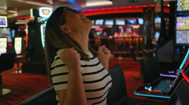 Neşeli genç bir yetişkin kadın, canlı bir geçmişi olan bir kumarhane kumar makinesinde kazandığı zaferi kutluyor.